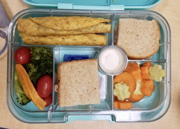 Bento lunch box with sandwich quarters, carrots and cucumbers in flower shapes, broccoli, tomato and ranch dip, and taquitos in separate compartments. 